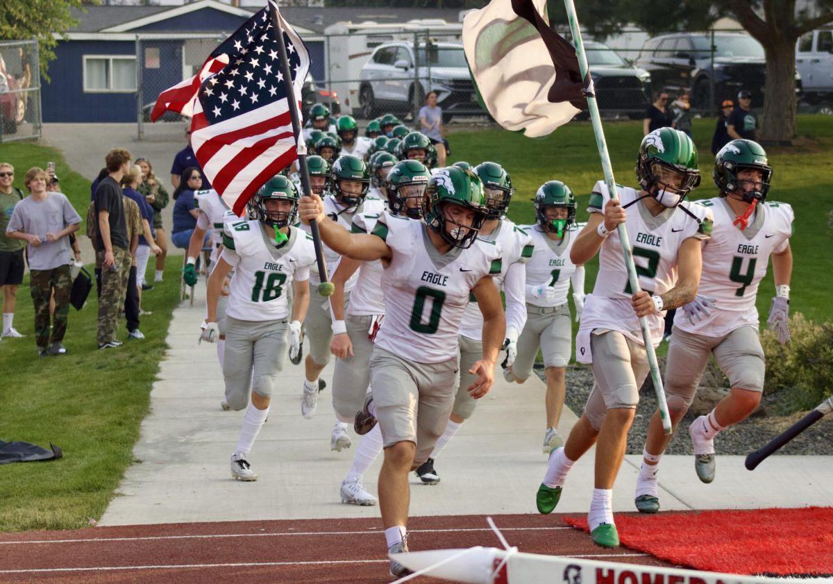 Senior Roman Walker and sophomore Austin Ramsey lead the football team as they run onto the field to play a football game. Walker is holding the American flag and Ramsey is hoist the Eagle High Stang Gang flag.